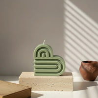 Bougie décorative en forme de cœur vert kaki, posée sur un socle, lumière naturelle.